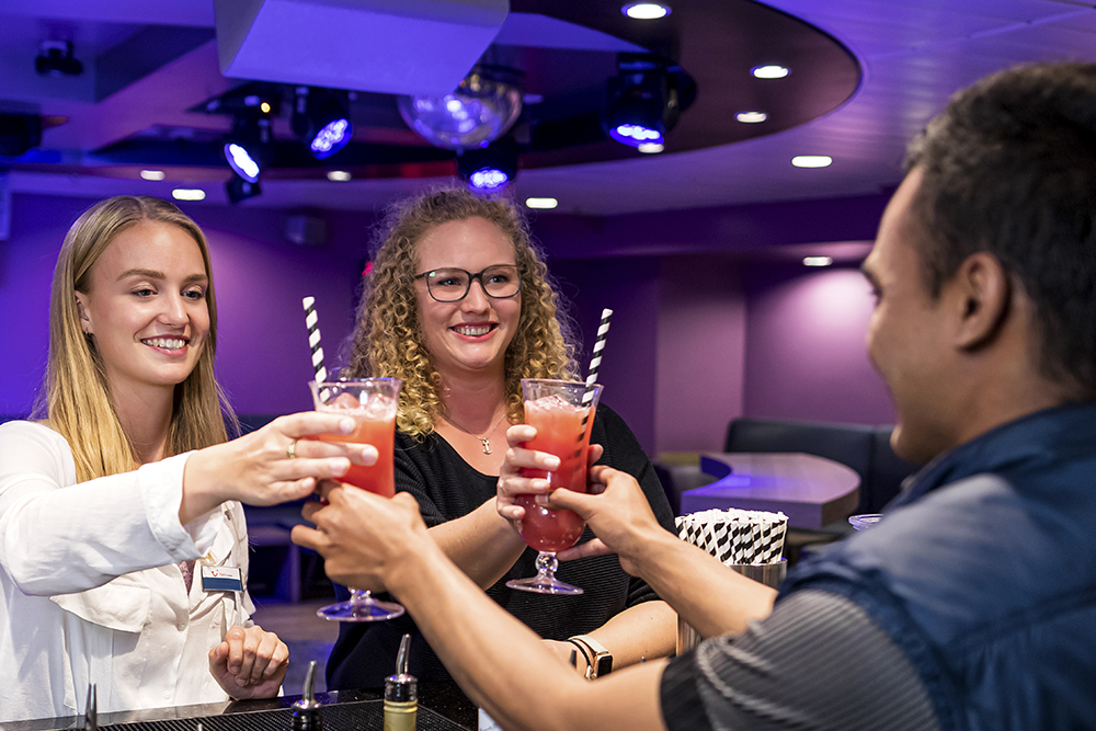 Zwei junge Crew Member stehen an einer Bar und bekommen von einem jungen Mann jeweils einen bunten Cocktail gereicht. 