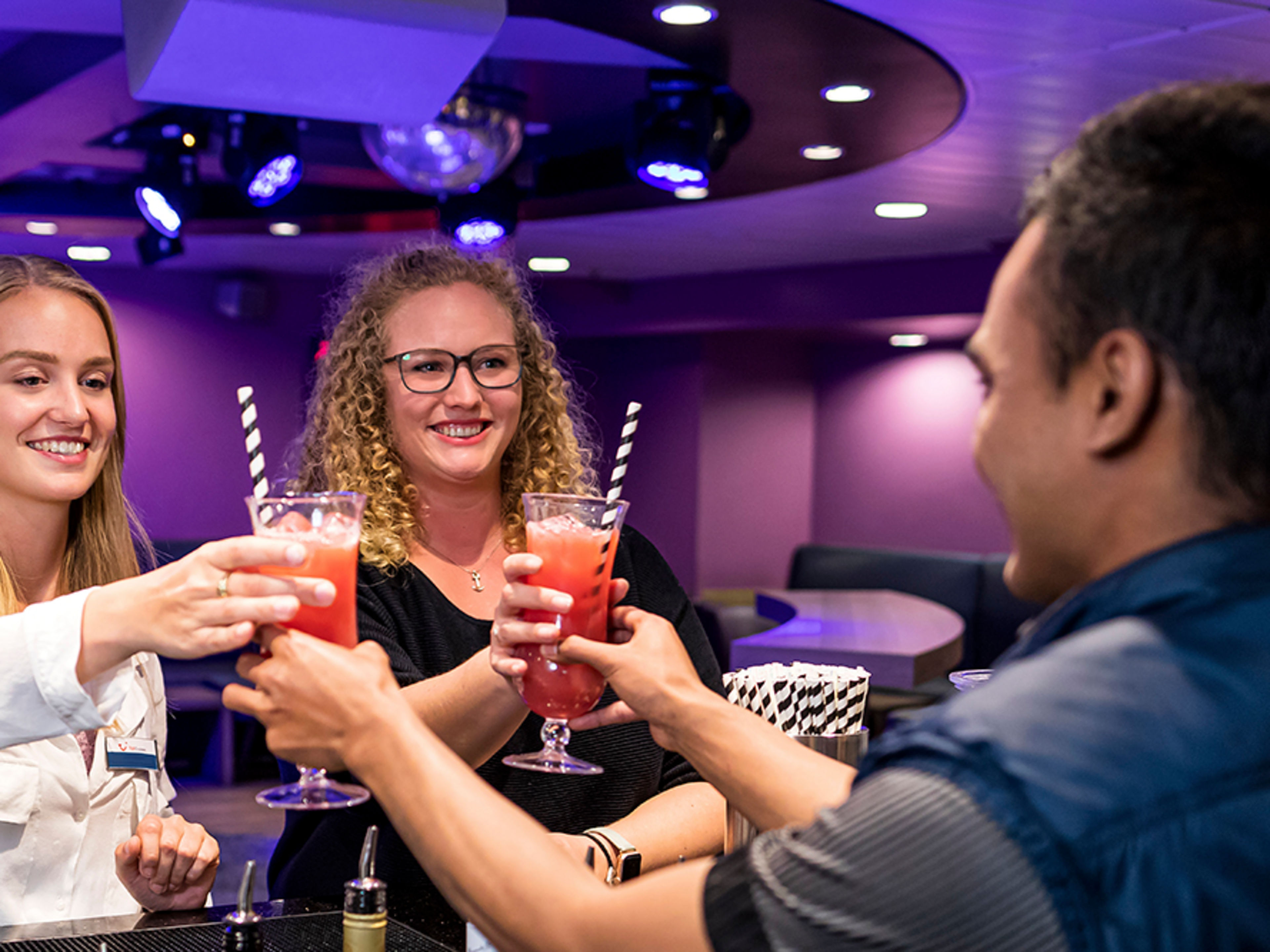 Zwei junge Crew Member stehen an einer Bar und bekommen von einem jungen Mann jeweils einen bunten Cocktail gereicht.