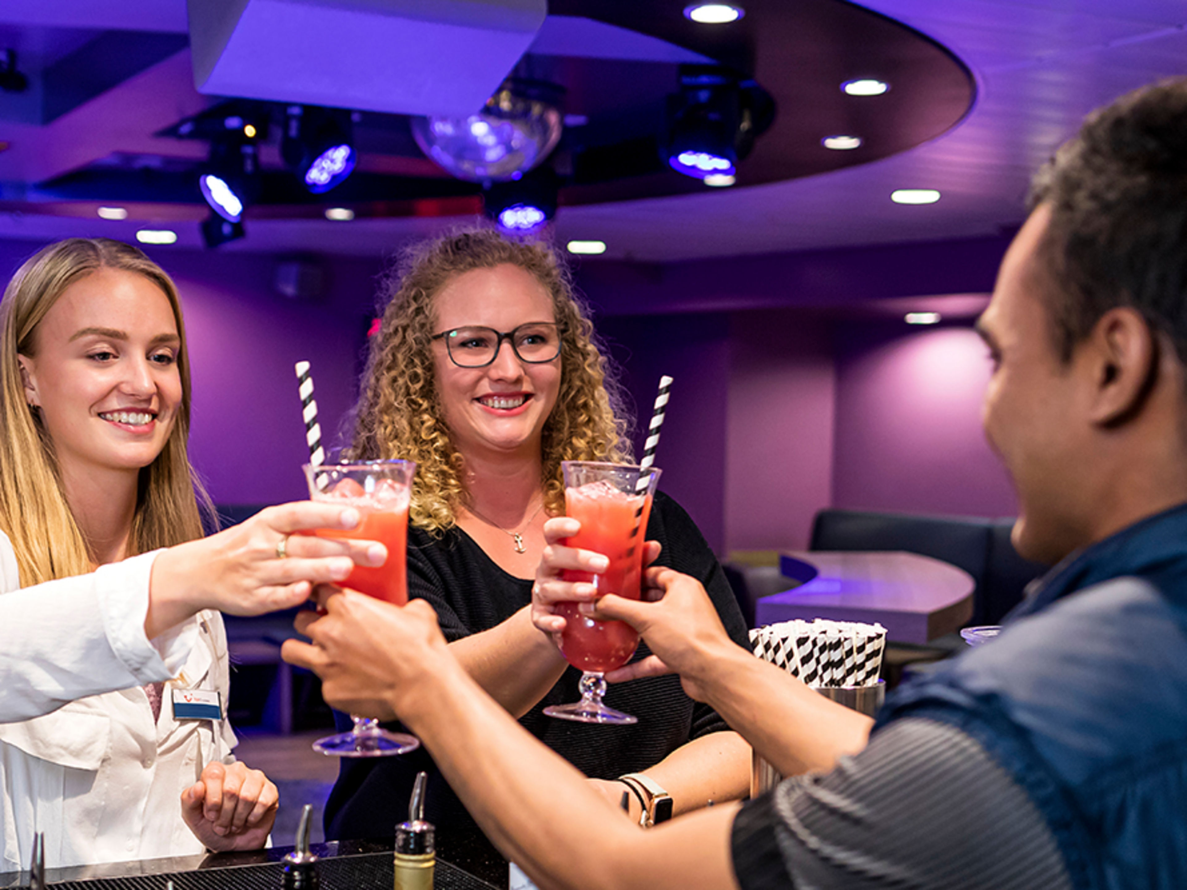 Zwei junge Crew Member stehen an einer Bar und bekommen von einem jungen Mann jeweils einen bunten Cocktail gereicht.