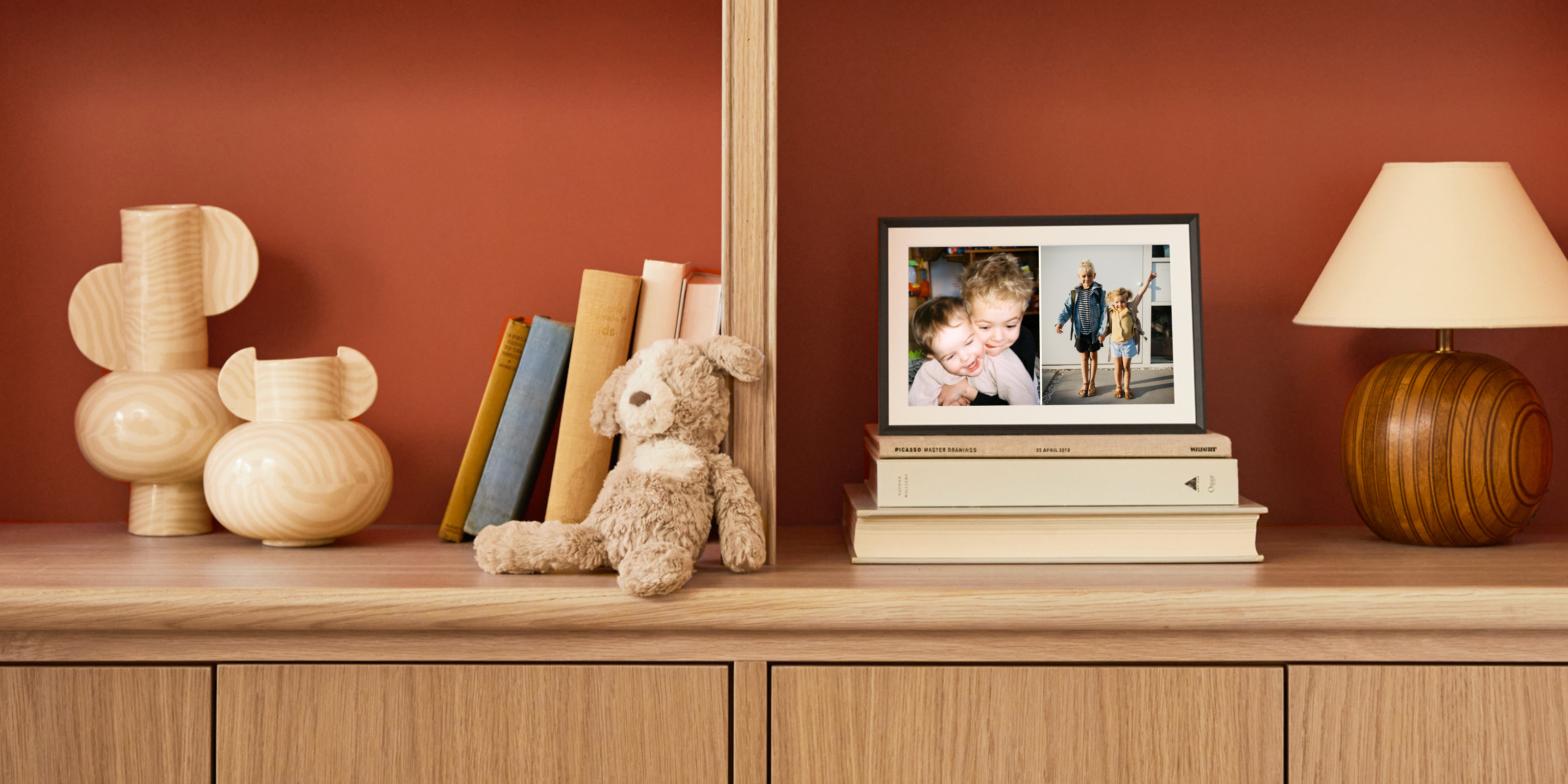 Aura digital frame on a shelf displaying two photos of young siblings, one hugging at home and one on the first day of school, beside books, a stuffed dog, and a lamp.