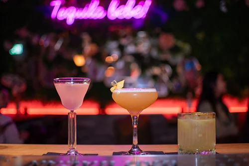 Three colorful cocktails on a bar, with a neon sign reading 'Tequila baby!'