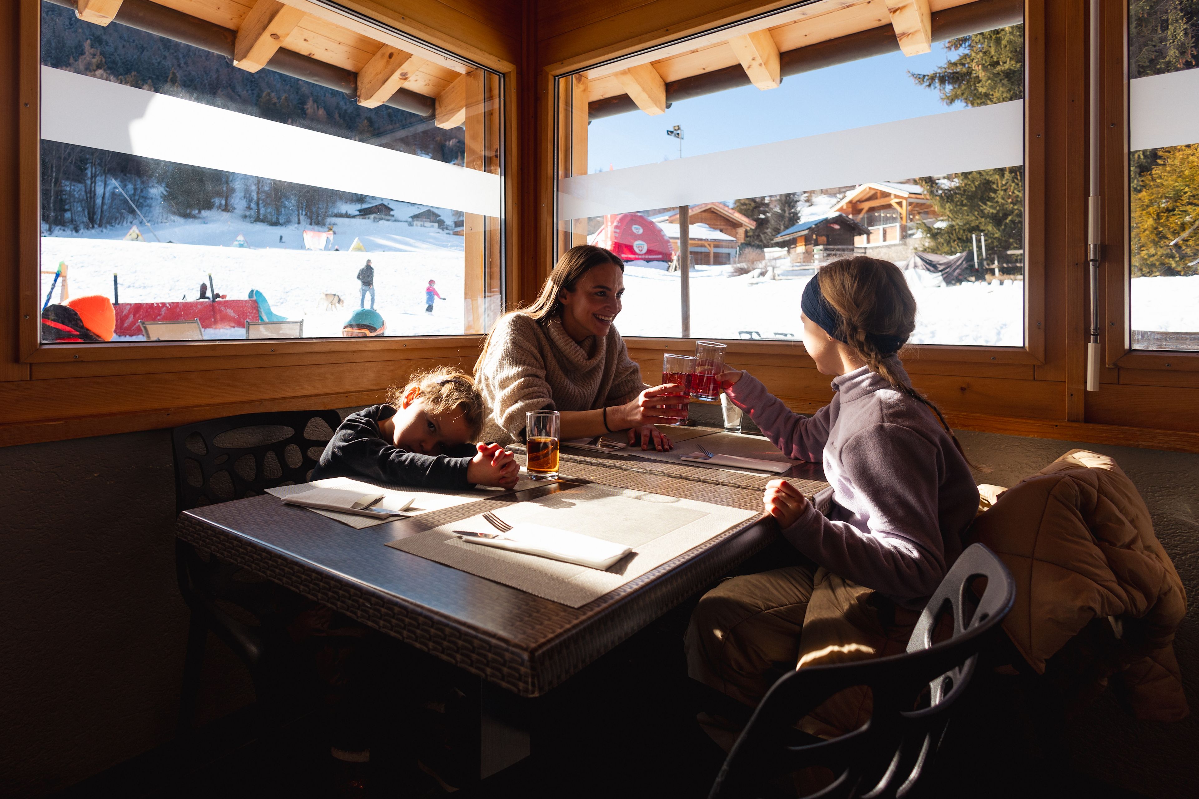 Eine Familie geniesst eine Pause im Restaurant am Fusse des Anfängerbereichs Le Lavioz in Vercorin, Wallis, mit Blick auf die verschneite Landschaft.