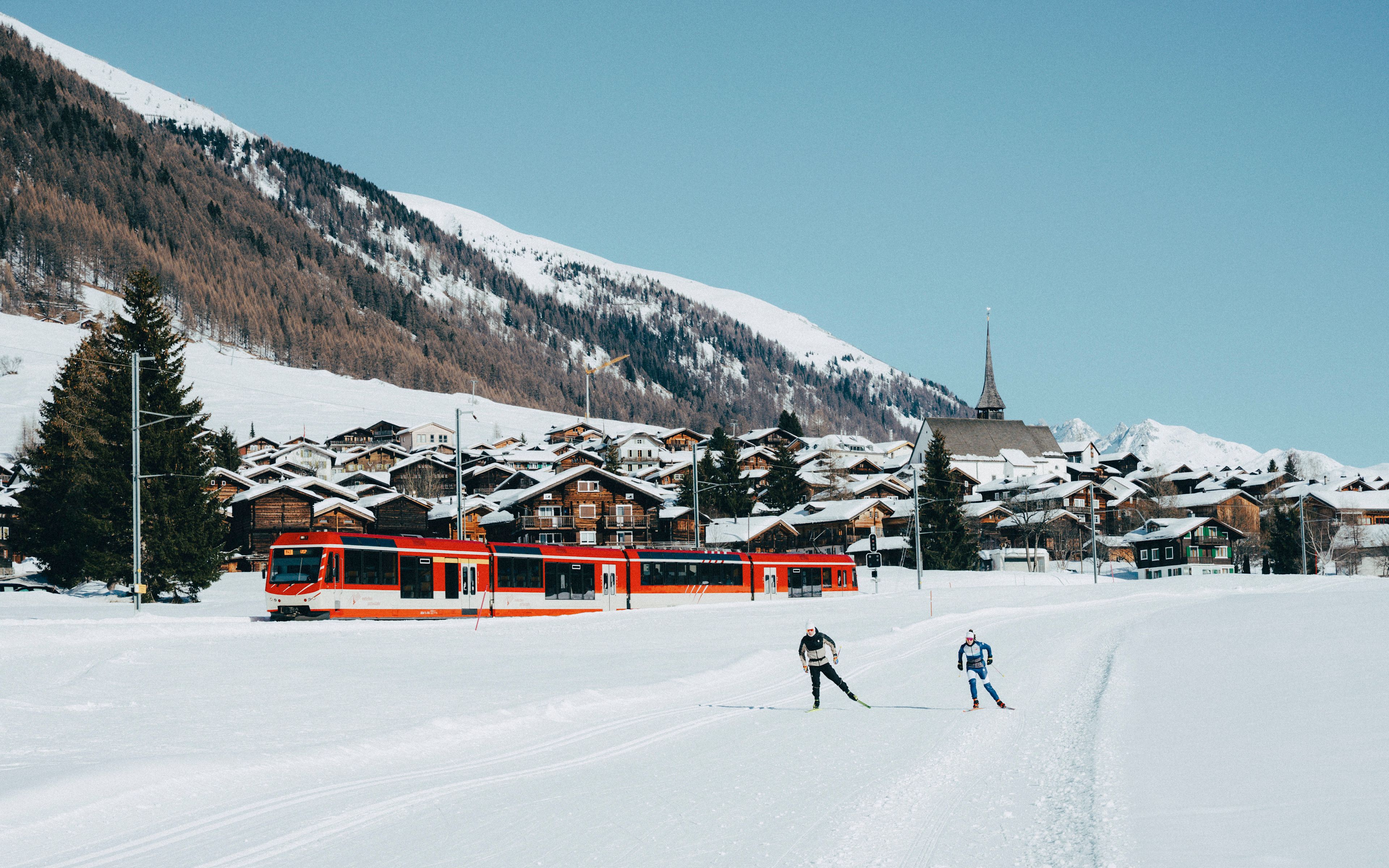 Two cross-country skiers in Goms with a passing train and snowy village in the background