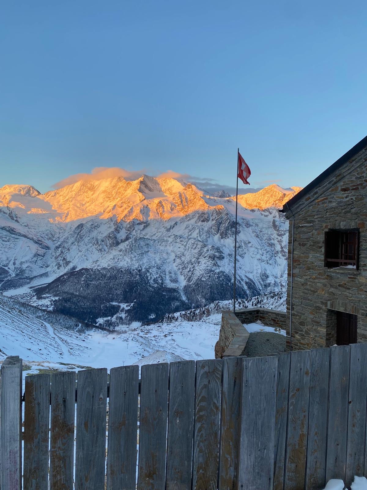 Blick auf die Weissmieshütte mit Schweizer Flagge und verschneiten Bergen im Sonnenaufgang in Saas-Grund