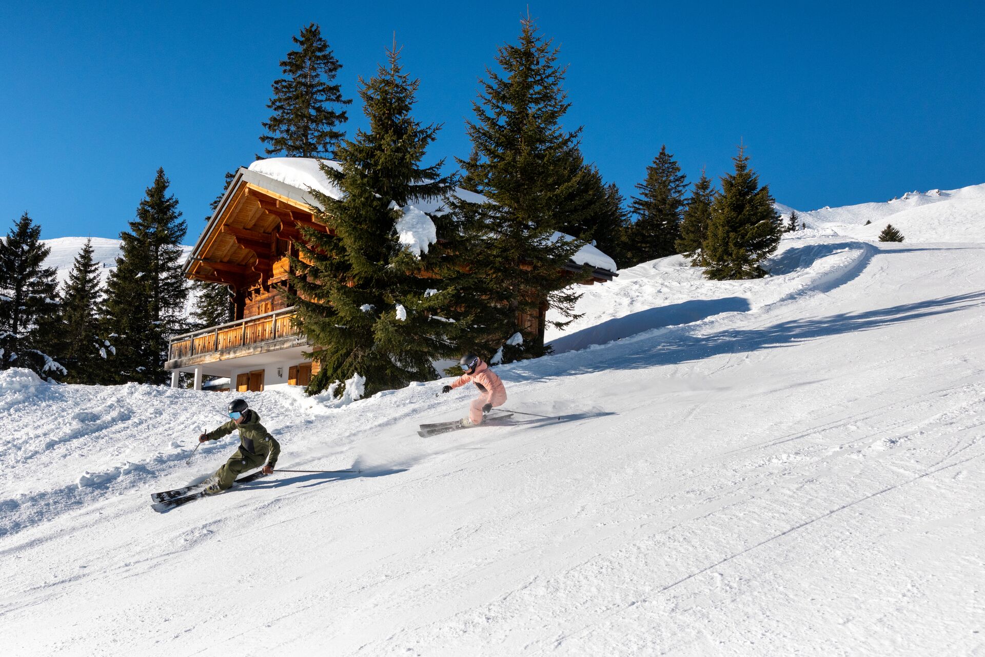 Deux skieurs dévalent une piste à Champéry, passant devant des sapins enneigés et un chalet en bois traditionnel