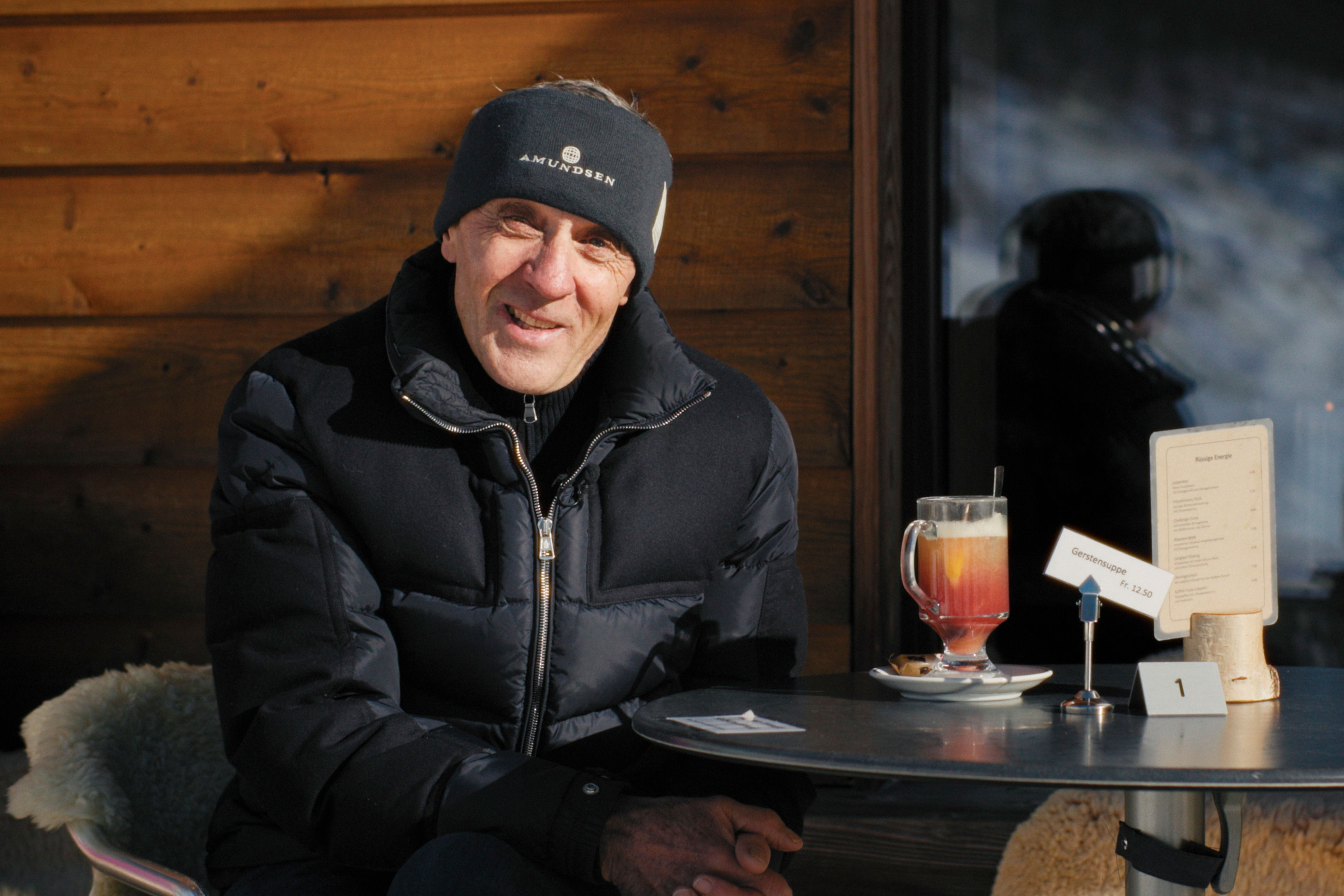Porträt von Koni Hallenbarter in Winterkleidung auf einer Sonnenterrasse in Goms, mit Getränk auf dem Tisch