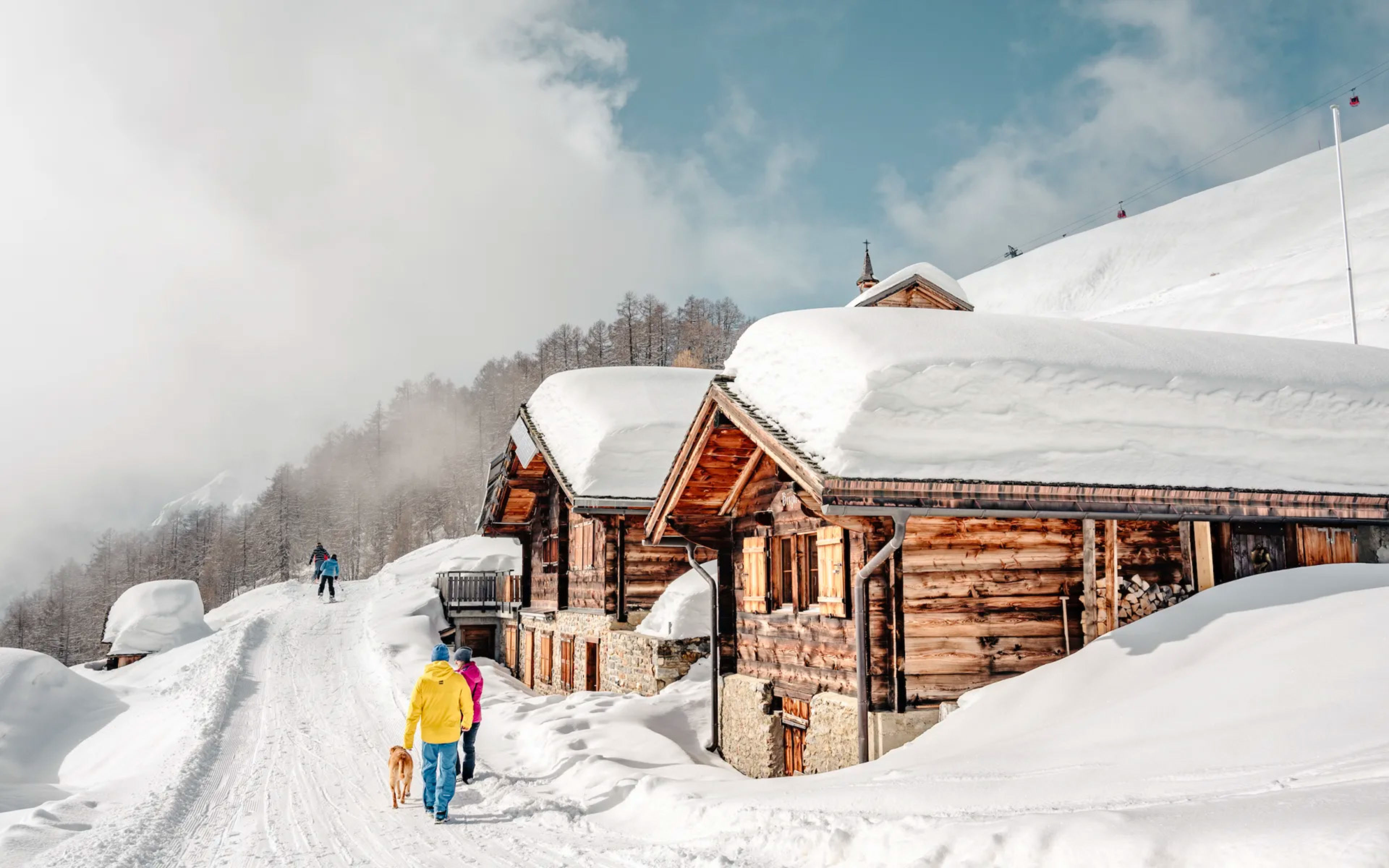 Un couple se promène dans la neige avec leur chien. Le chemin longue des chalets enneigés à Torrent, Loèche-les-Bains. Valais, Suisse