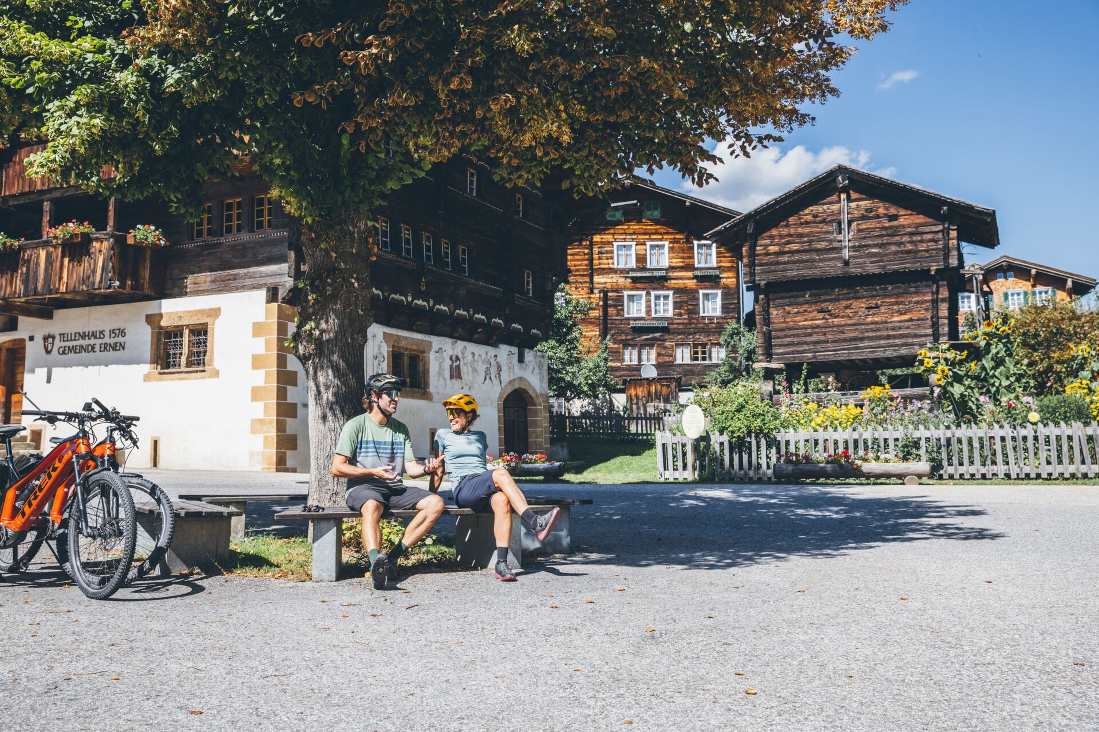 Two mountain bikers relaxing in the shade in front of the Tellenhaus in the village center of Ernen with traditional wooden houses, Valais, Switzerland