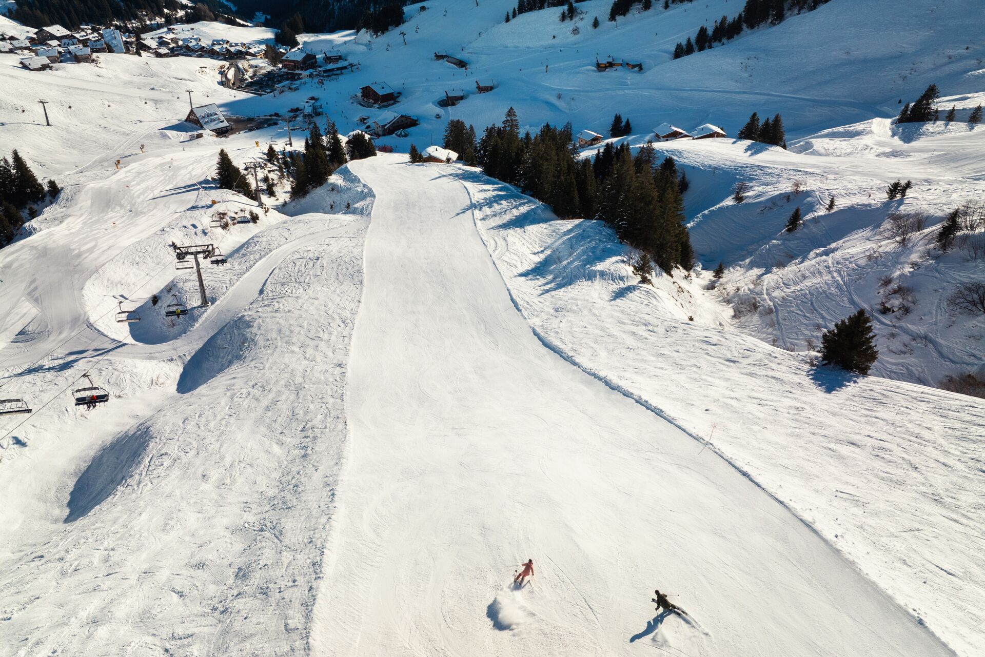 Vue drone d’une large piste de ski à Champéry avec deux skieurs, entourée de chalets et de sapins