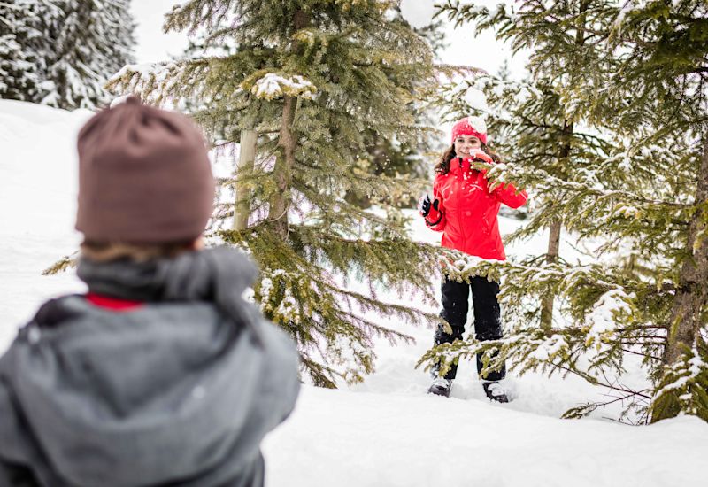 Un frère et sa soeur font une bataille de boule de neige. La soeur se cache derrière les arbres enneigés. Valais, Suisse