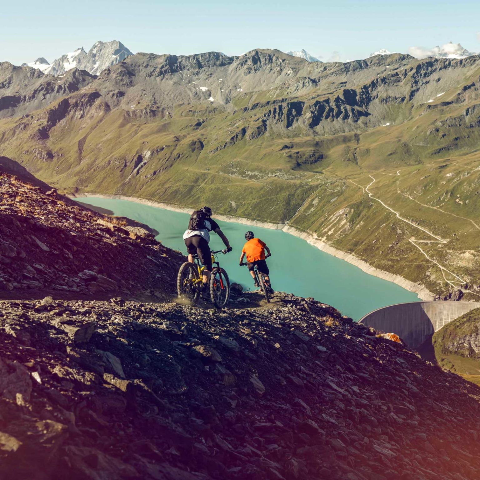 Two mountain bikers in the descent of a mountain bike route with the view on the lake of Moiry. Valais, Switzerland