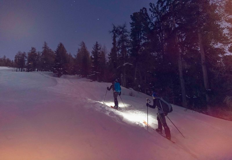 Deux personnes font du ski de randonnée nocturne, éclairé à l'aide de leur lampe frontale et la lune. Valais, Suisse