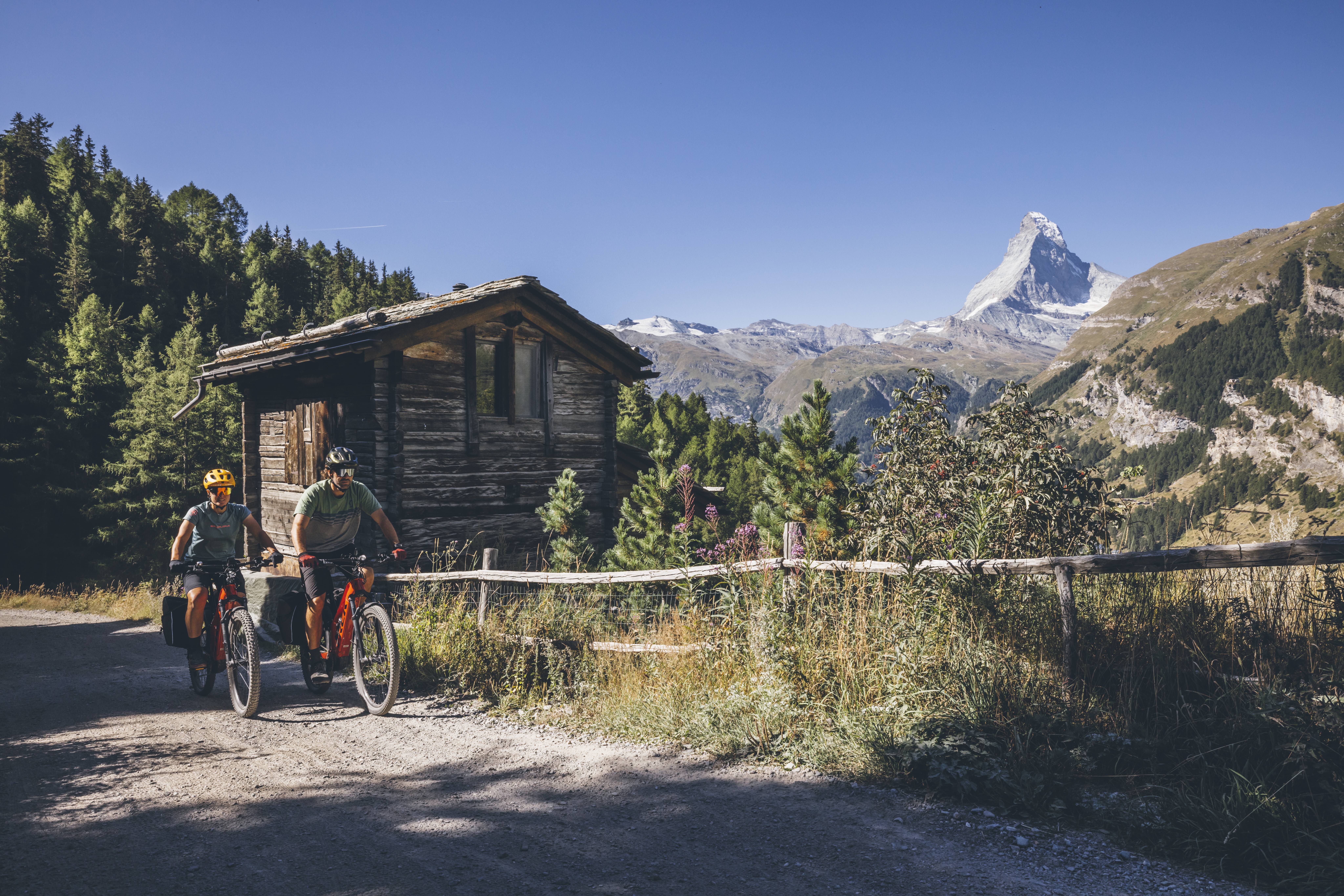 Two mountain bikers on a gravel road in the Swiss Alps with a view of the Matterhorn, following the Glacier Bike Tour West route, Valais, Switzerland