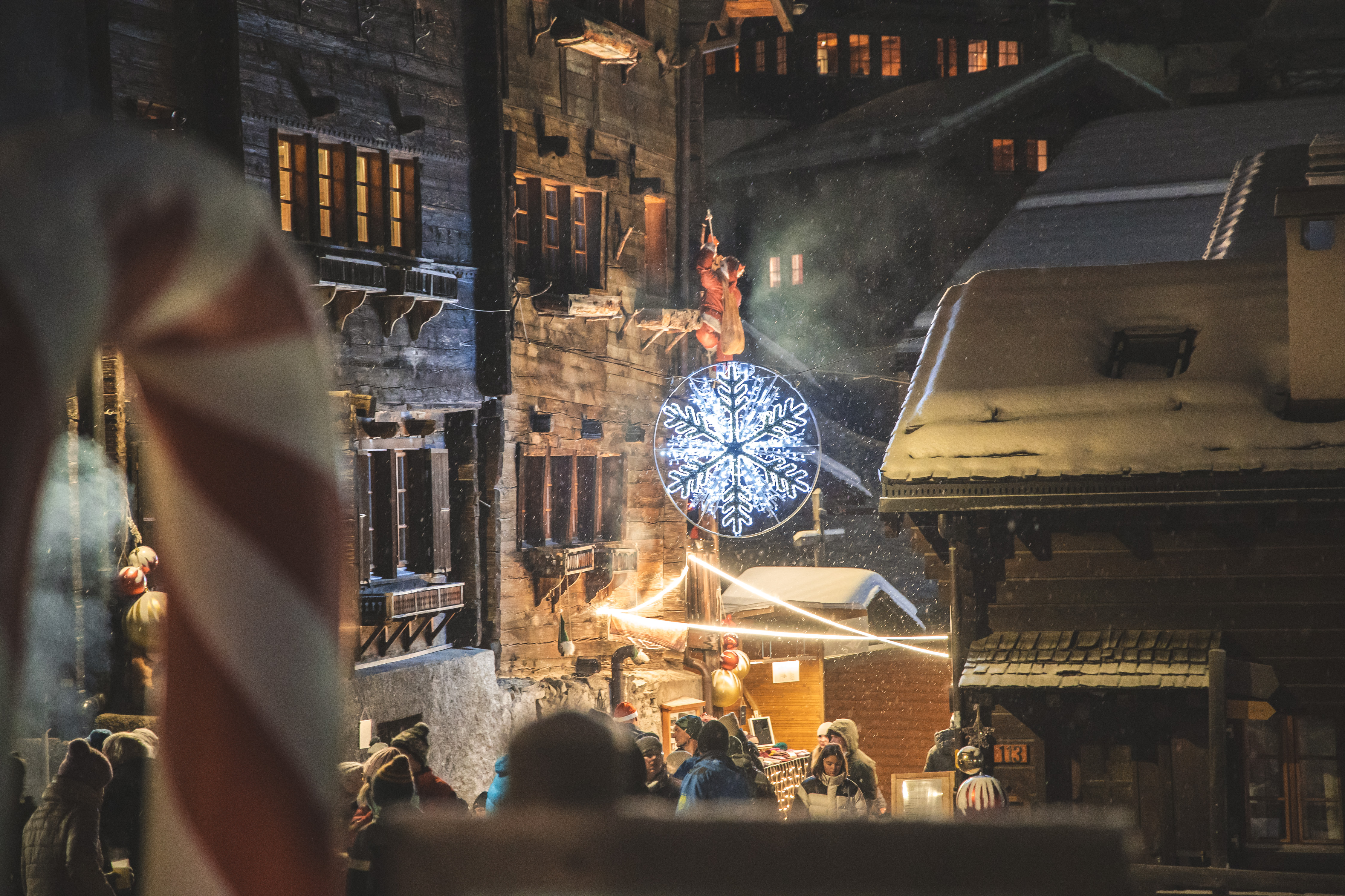 Menschen schlendern über den verschneiten Weihnachtsmarkt in Grimentz, umgeben von Holzhäusern und Weihnachtsbeleuchtung