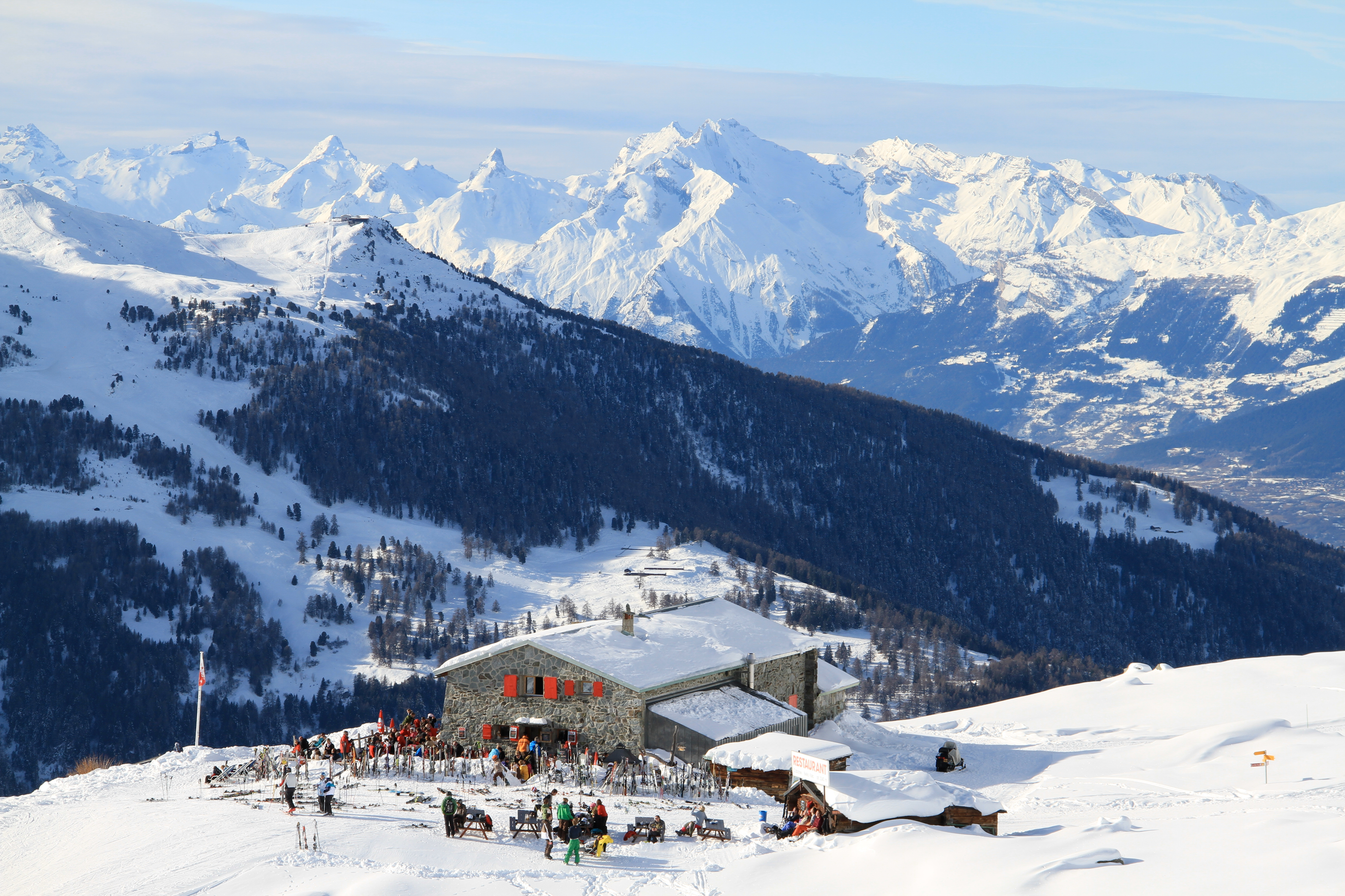Restaurant de la Cabane Bella-Tola à St-Luc, Valais, entouré de montagnes enneigées avec terrasse ensoleillée et skieurs