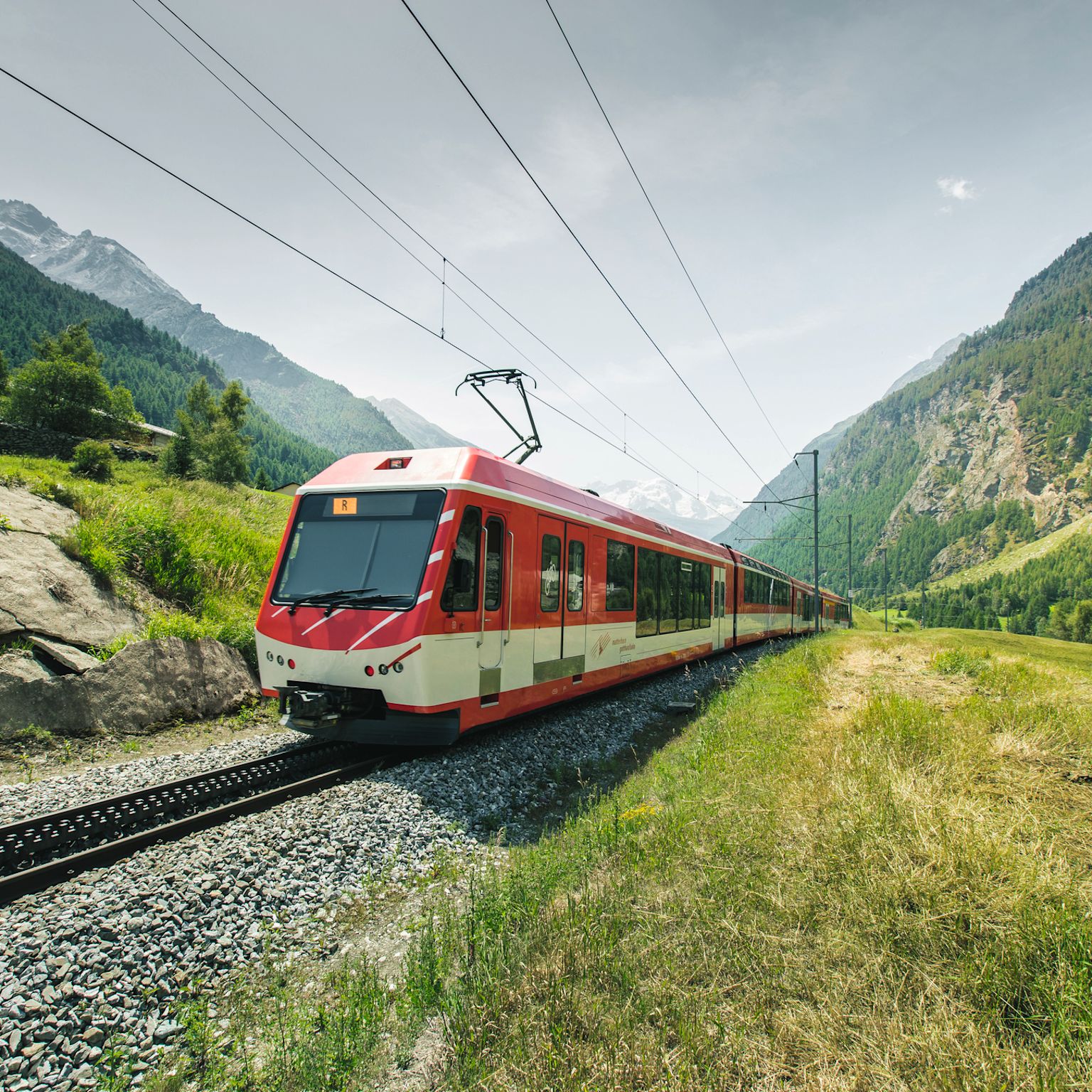 Roter Zug der Matterhorn Gotthard Bahn bei der Durchfahrt durch das Dorf Randa im Kanton Wallis, mit Blick auf die umliegenden Berge