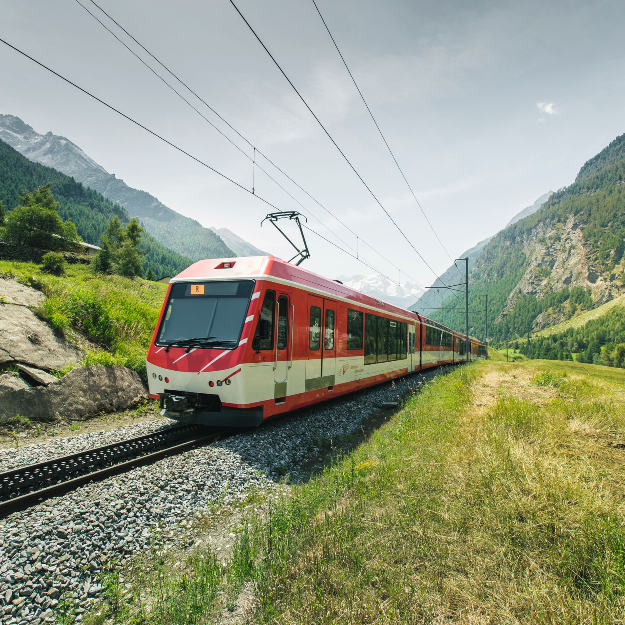 Roter Zug der Matterhorn Gotthard Bahn bei der Durchfahrt durch das Dorf Randa im Kanton Wallis, mit Blick auf die umliegenden Berge