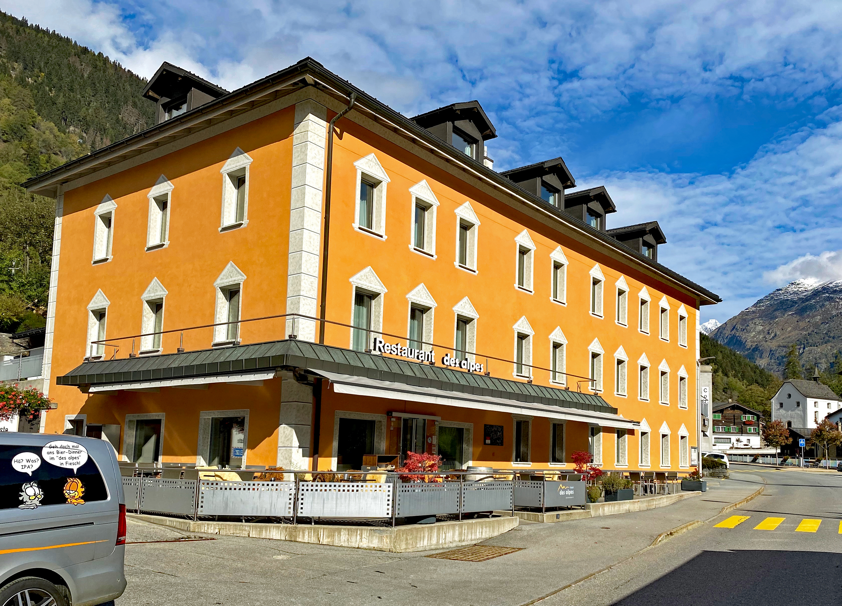 Außenansicht des orangefarbenen Hotels des Alpes mit Terrasse im Dorfzentrum von Fiesch