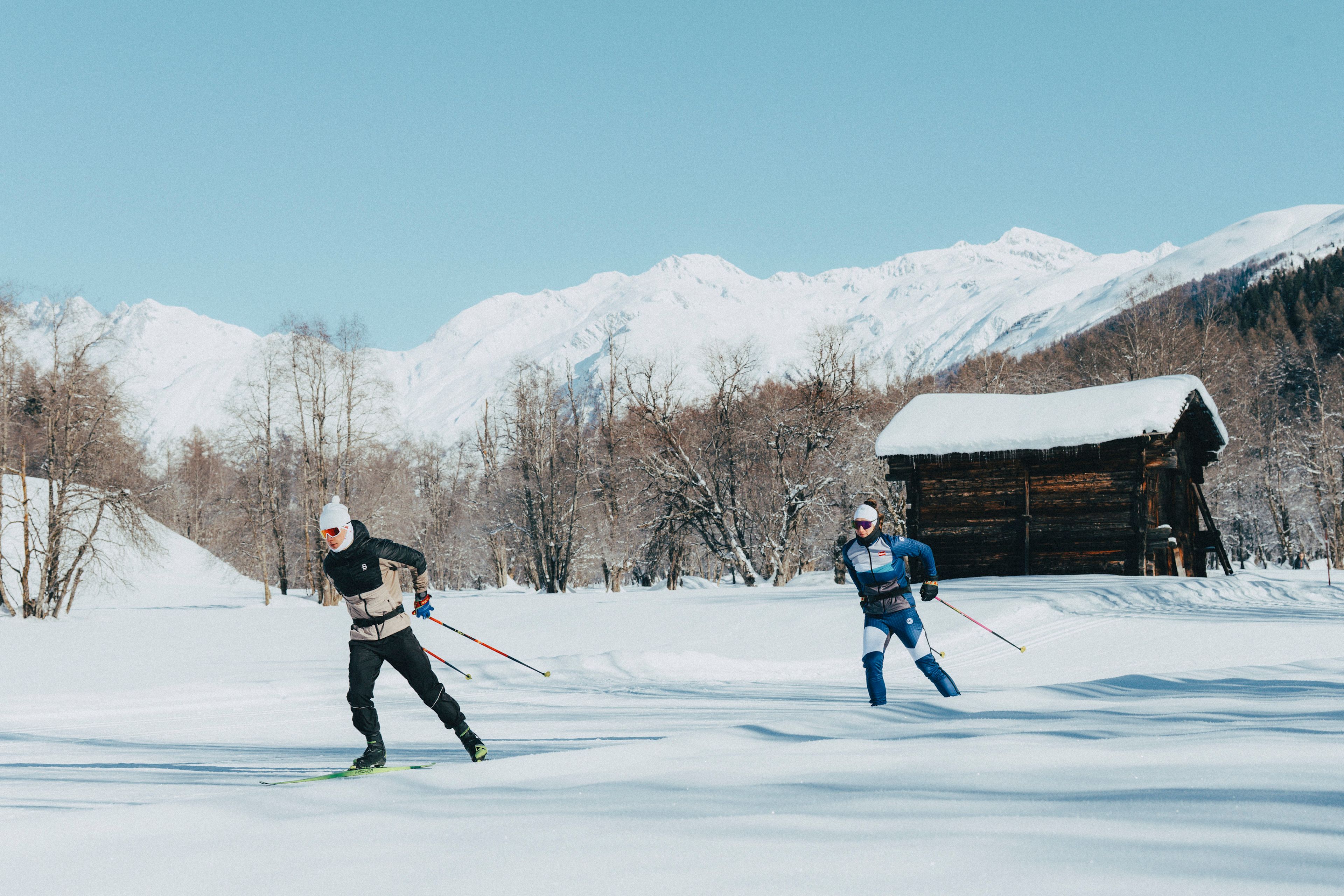 Two cross-country skiers on a snowy trail in the winter landscape of Goms with a wooden hut in the background