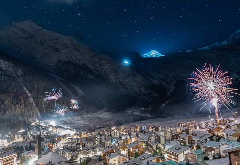 Feu d’artifice nocturne à Saas-Fee au-dessus des toits enneigés, avec les montagnes en toile de fond et une ambiance festive de Nouvel An