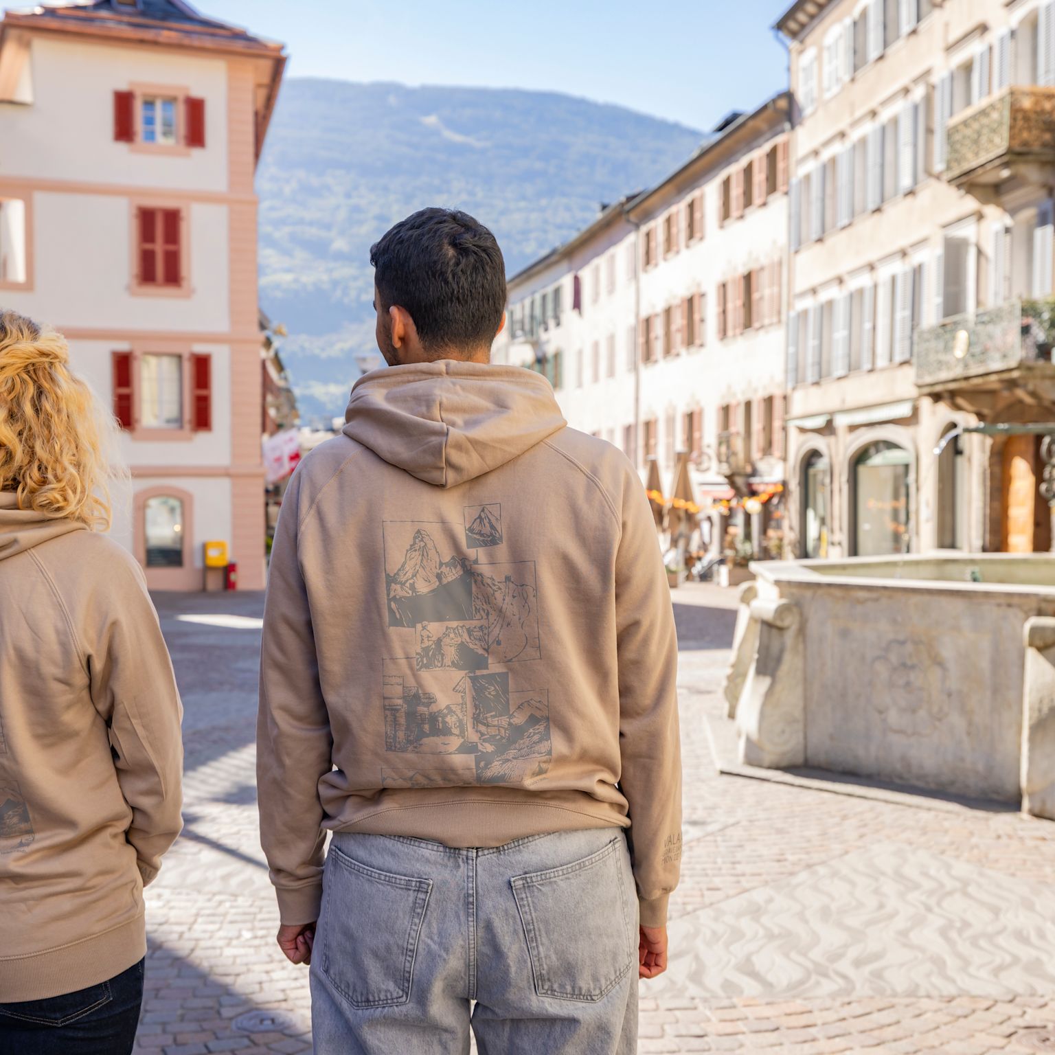 Rückansicht zweier Personen mit beigem Hoodie “Valais Horizons” in der Altstadt von Sion