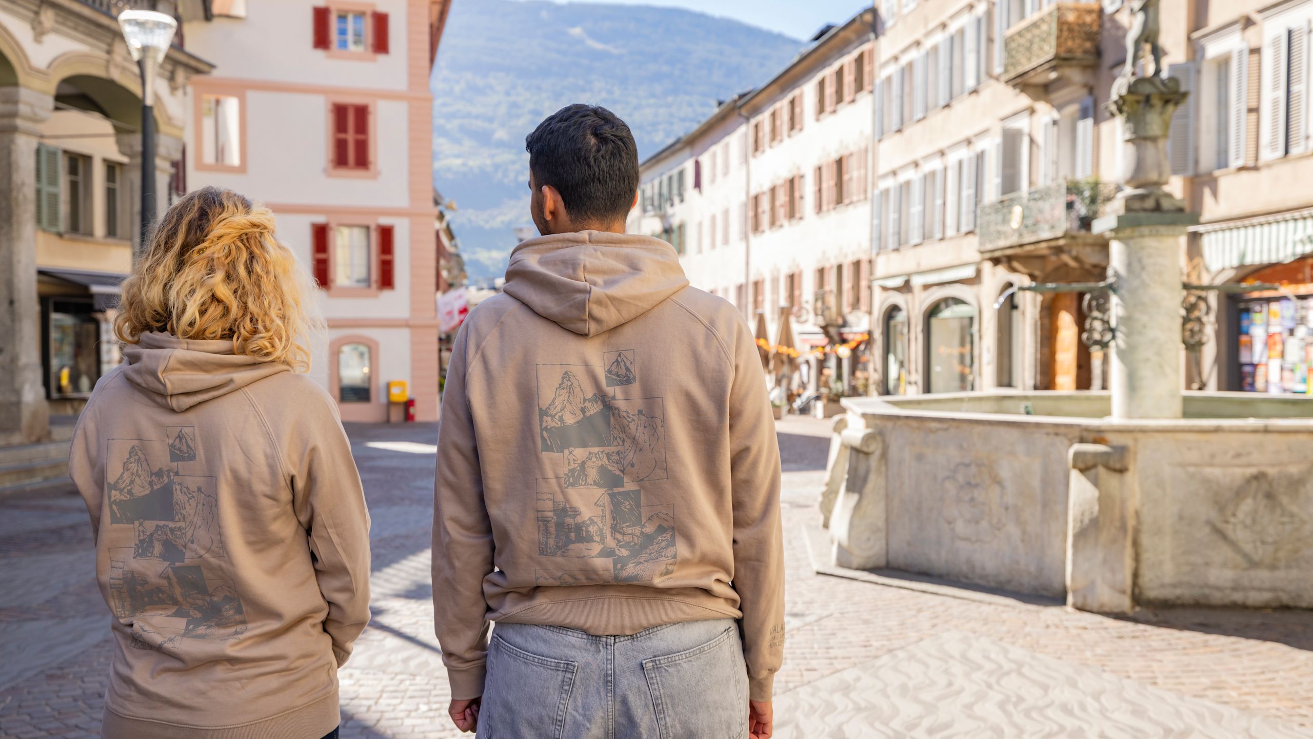 Rückansicht zweier Personen mit beigem Hoodie “Valais Horizons” in der Altstadt von Sion