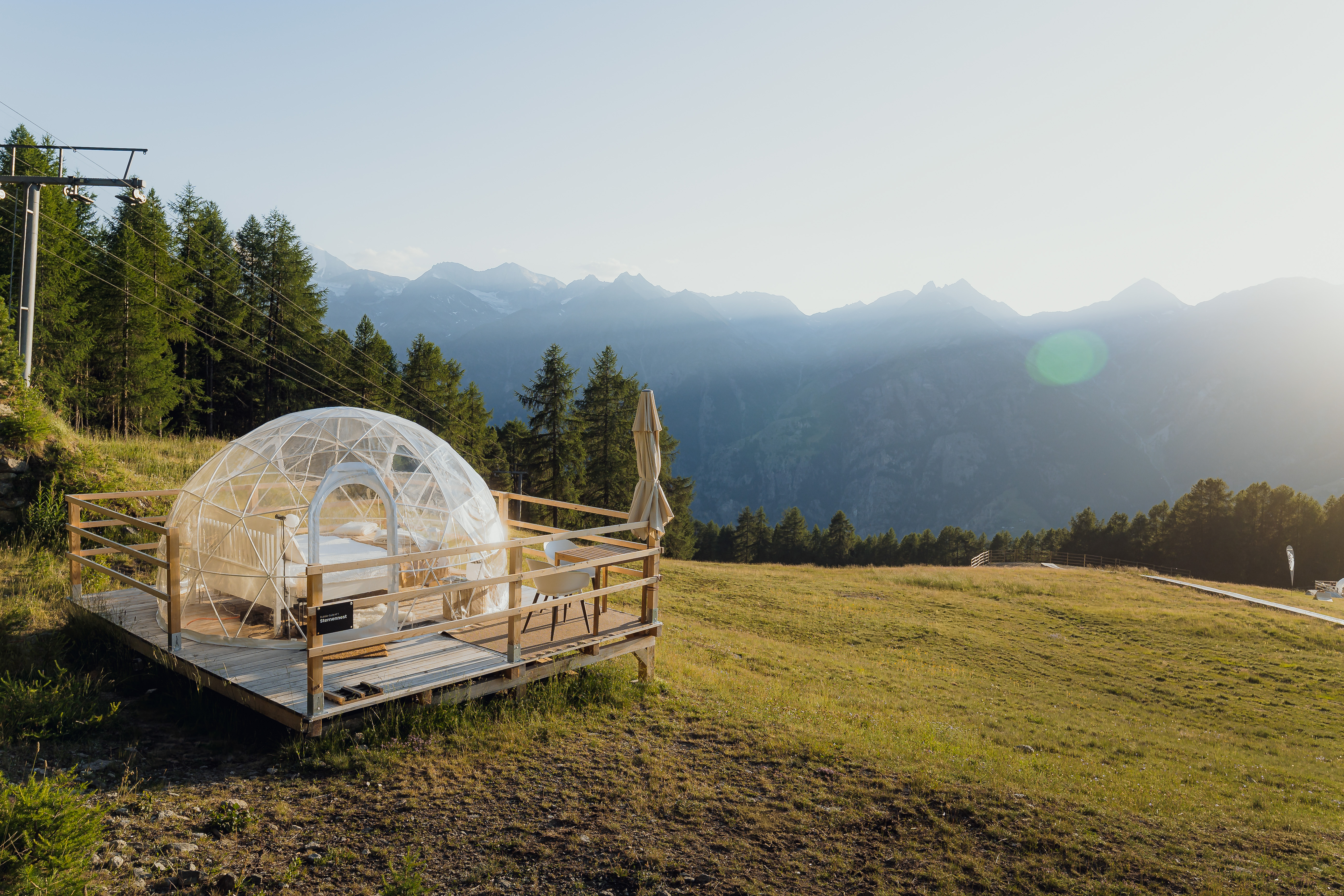 Transparent geodesic dome tent on wooden deck in alpine meadow with mountain range backdrop and ski lift visible.