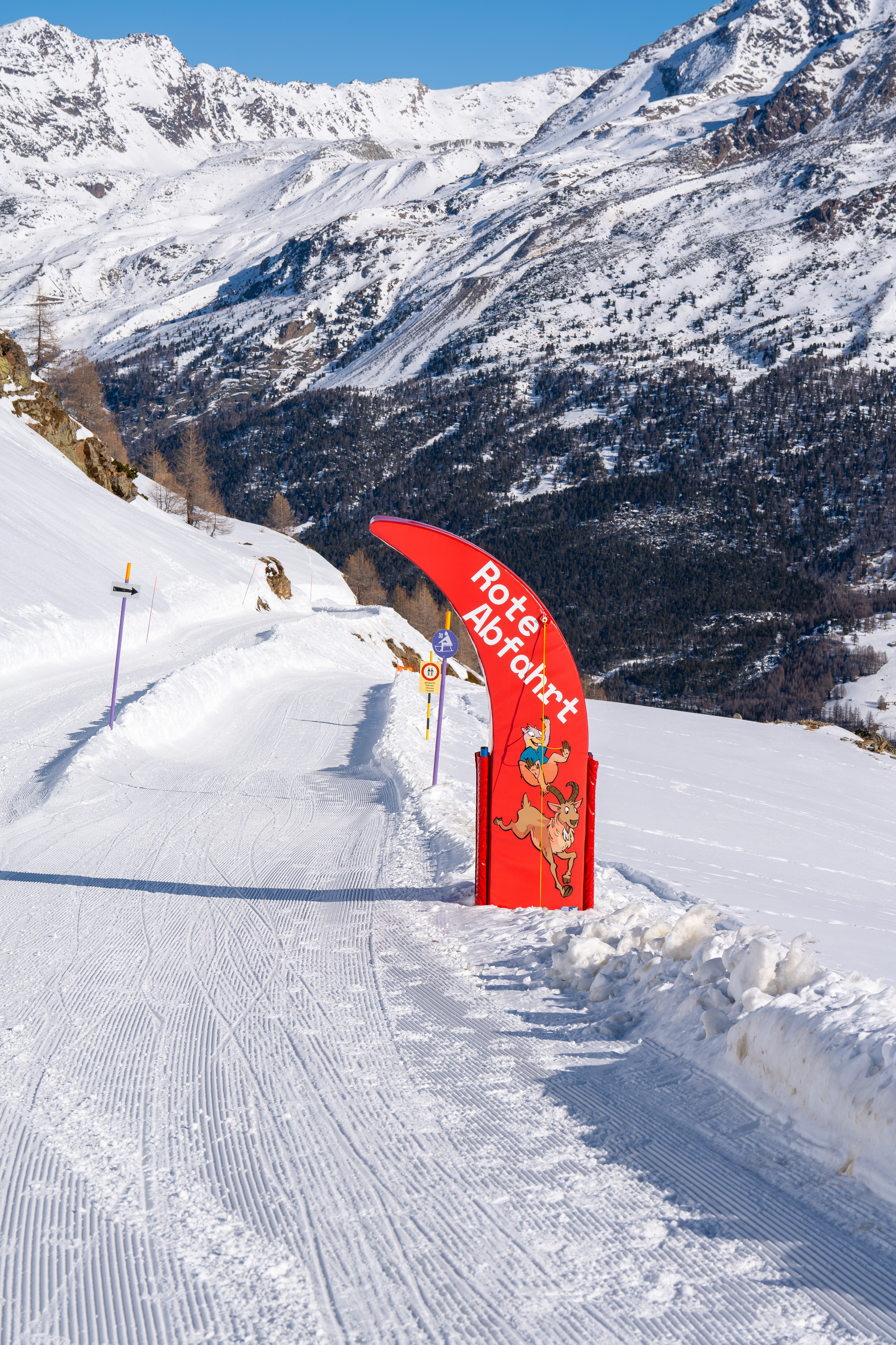 View of the red sledding run Hannig in Saas-Fee, Valais, with a signpost and snowy mountains in the background.