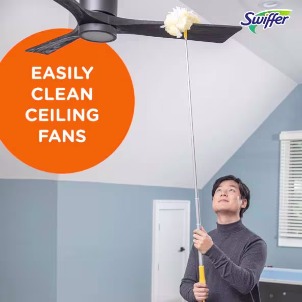 A person using a Swiffer to clean a ceiling fan in a bright room with light blue walls and white trim