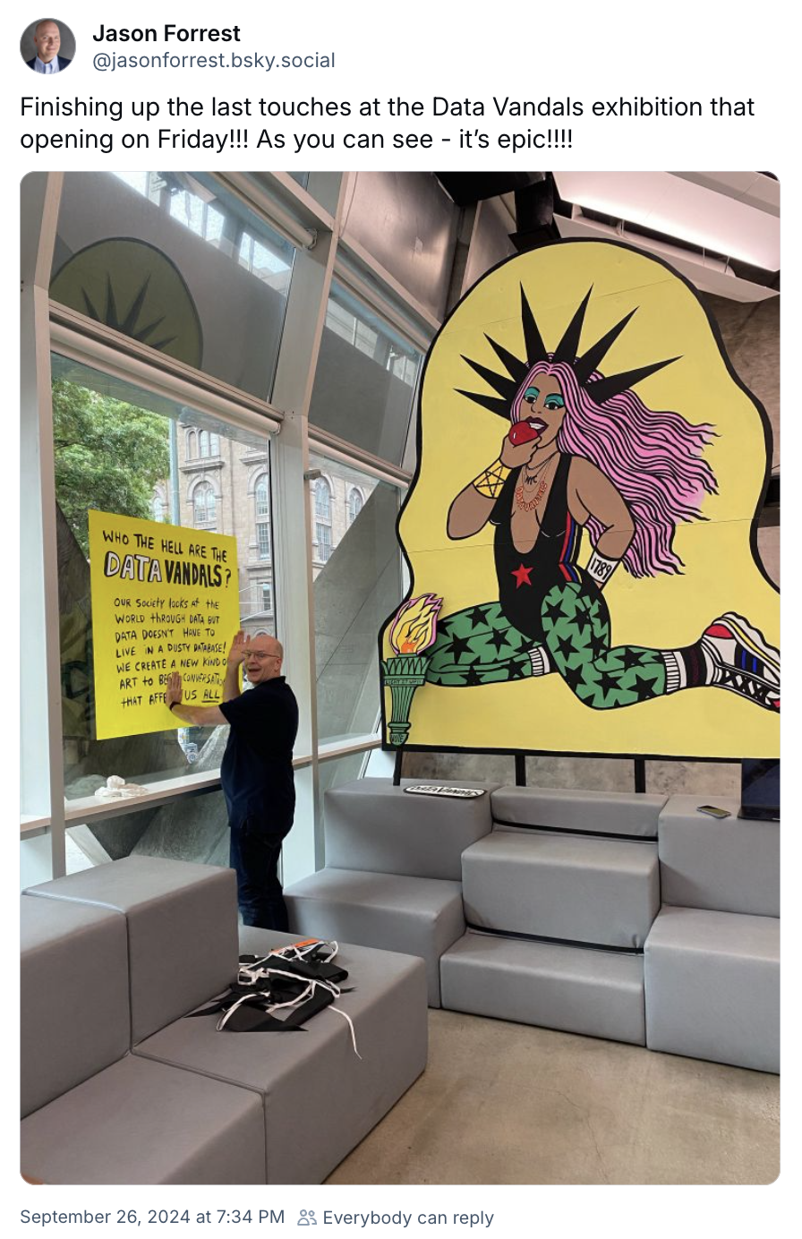 Jason Forrest hangs a yellow poster for the Hello from the Data Vandals show at Cooper Union, New York City. In the background is a large pop art painting of a woman with long pink hair and wearing green tights with starts on them, over a yellow background