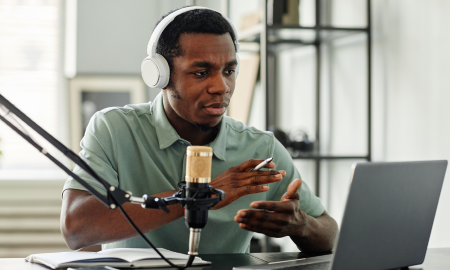 An individual in headphones is sitting at a desk speaking into a microphone and looking at a laptop, evidently engaged in a podcast or online interview.
