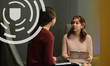 Two individuals in a discussion at a table, one holding a tablet. Both are seated in a modern office setting with a neutral background.