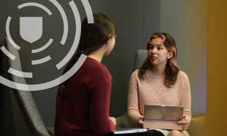 Two individuals in a discussion at a table, one holding a tablet. Both are seated in a modern office setting with a neutral background.