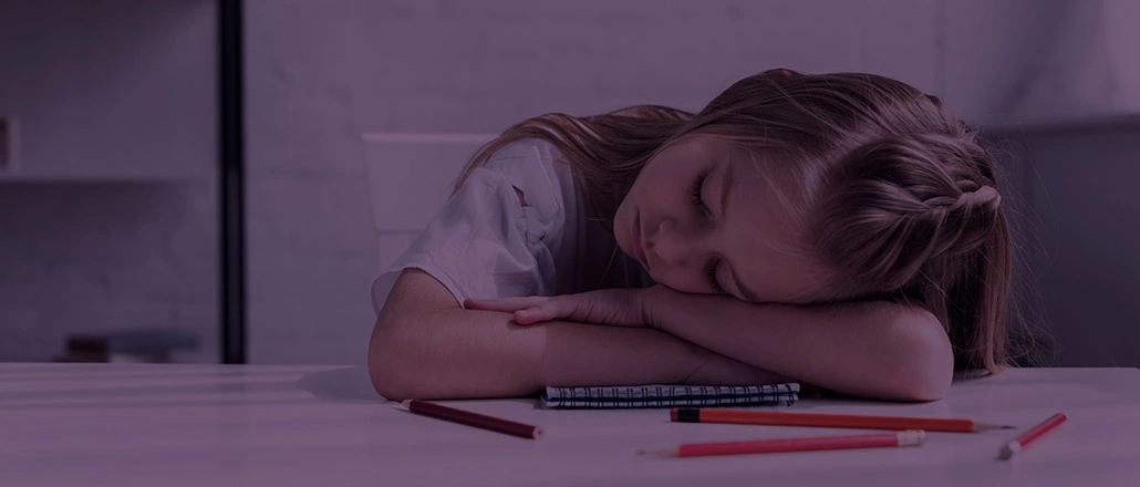 “Kid resting her head on a table with colored pencils.”