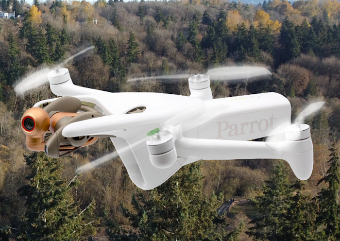 A white Parrot drone is flying over a forested landscape. The drone has four propellers and a camera mounted on the front, capturing the surrounding trees against a backdrop of a clear sky.