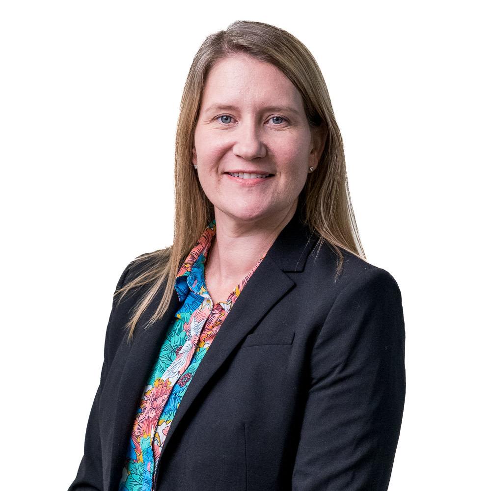 Smiling woman with long blonde hair wearing a gray blazer over a dark floral blouse, standing in front of a white wall with a grid pattern.
