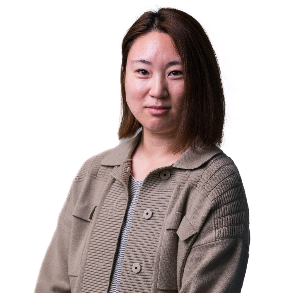 A woman with long dark hair and a friendly smile stands in front of a wall with a patterned design. She is wearing a white shirt and a gray cardigan.
