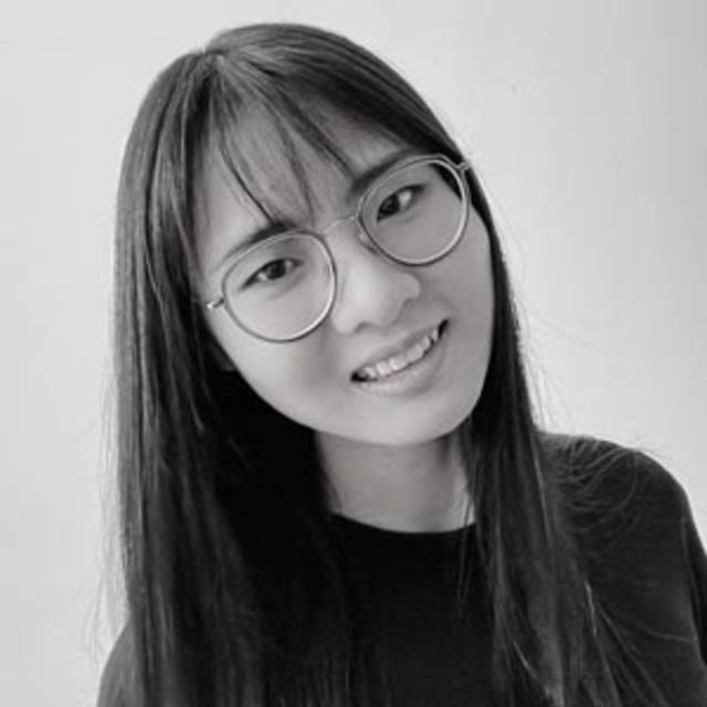 A person with long, straight hair is wearing round glasses and a black shirt, smiling slightly. The background is plain and light-colored. The image is in black and white.