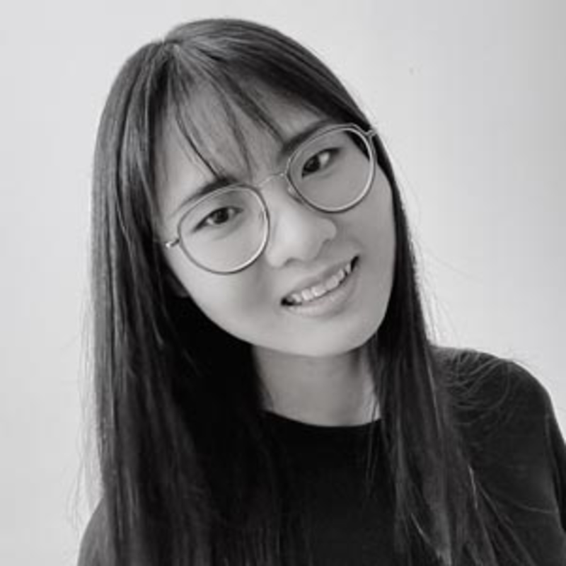 A person with long, straight hair is wearing round glasses and a black shirt, smiling slightly. The background is plain and light-colored. The image is in black and white.