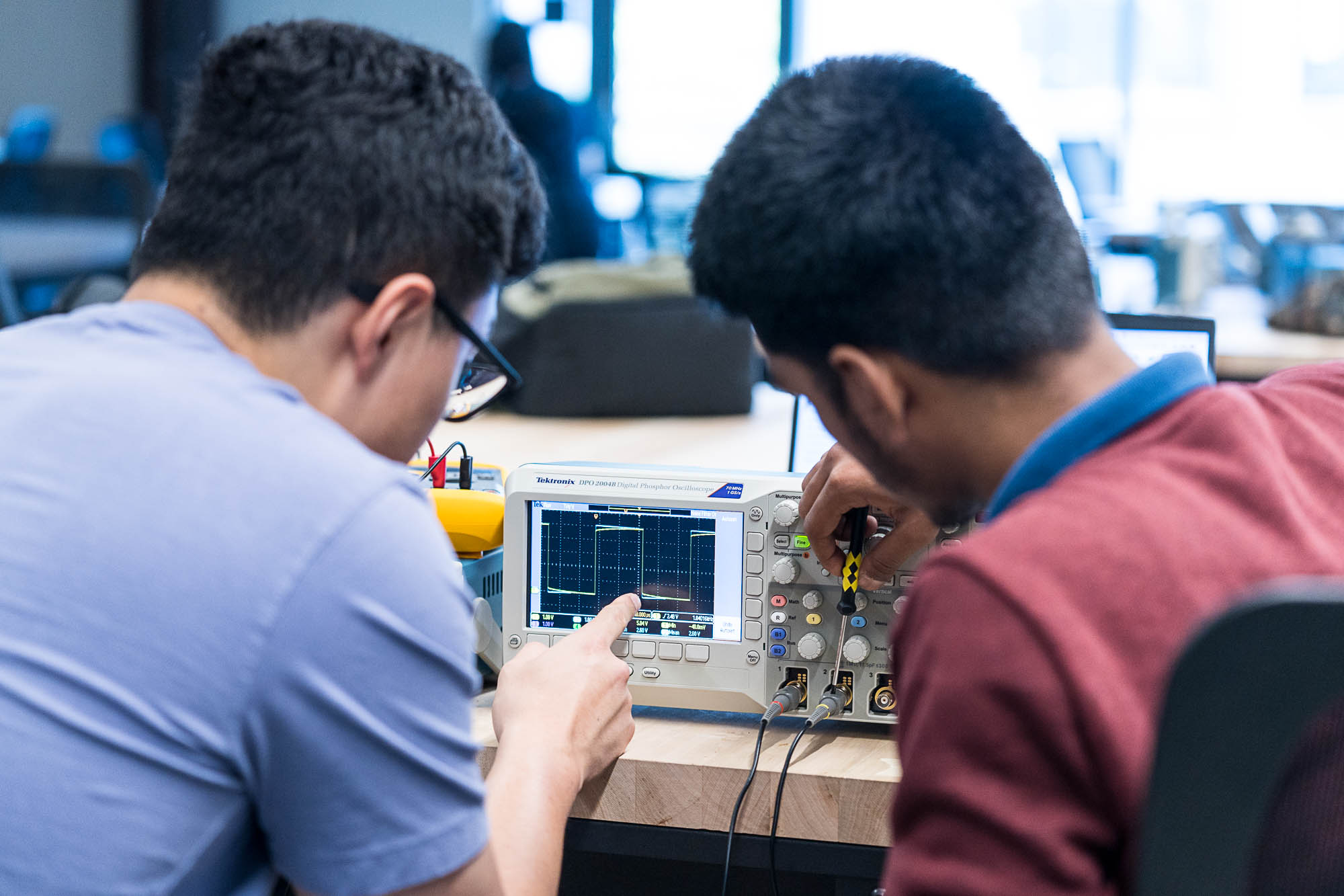 Two individuals are using an oscilloscope on a desk. One person points at the screen while the other adjusts the devices controls. The setting appears to be a classroom or lab with a focus on electronics work.