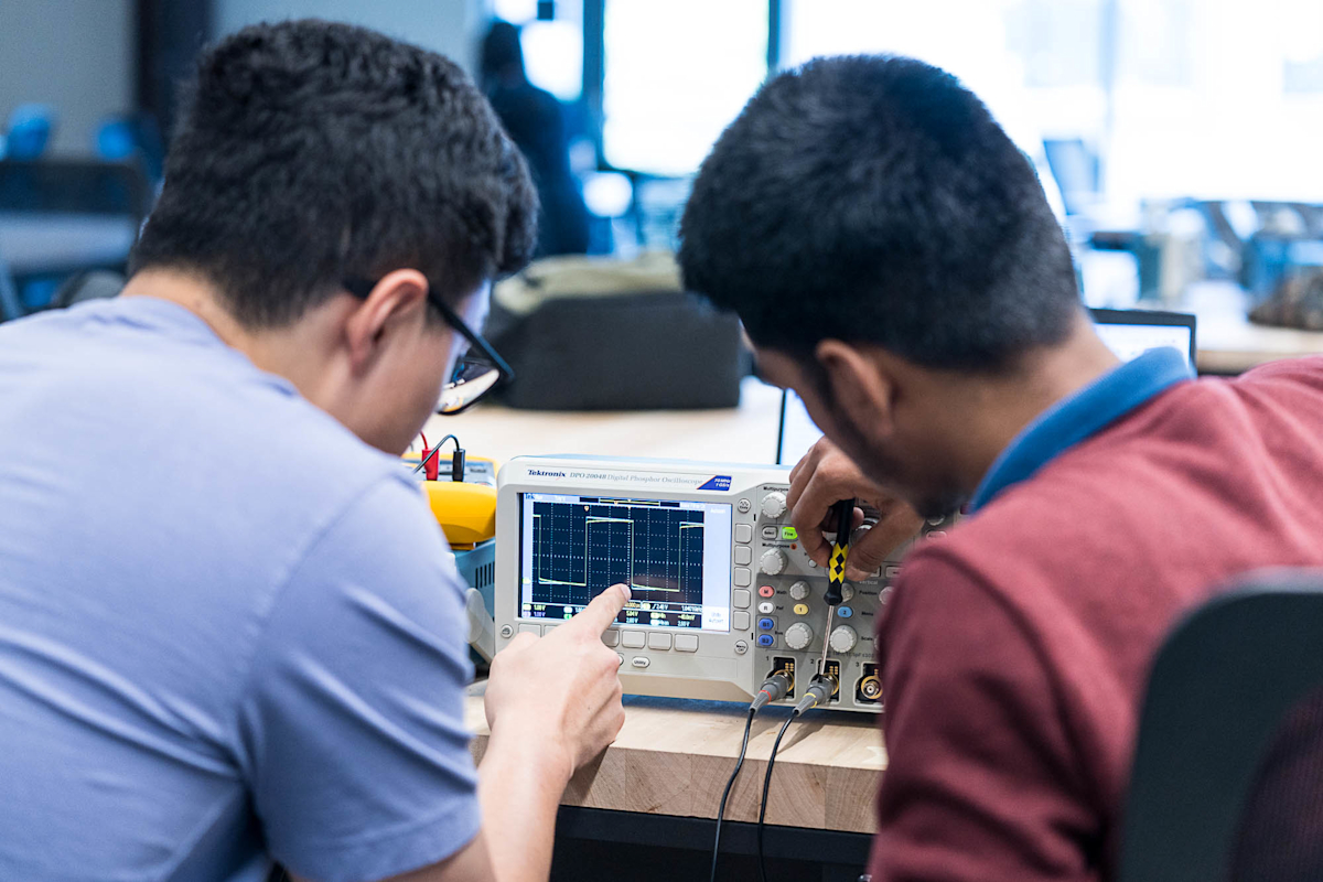 Two individuals are using an oscilloscope on a desk. One person points at the screen while the other adjusts the devices controls. The setting appears to be a classroom or lab with a focus on electronics work.