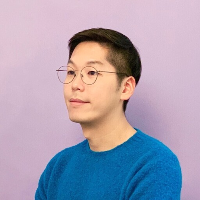 A person with short black hair and round glasses is wearing a bright blue sweater. They are looking slightly upward to the side against a plain lavender background.