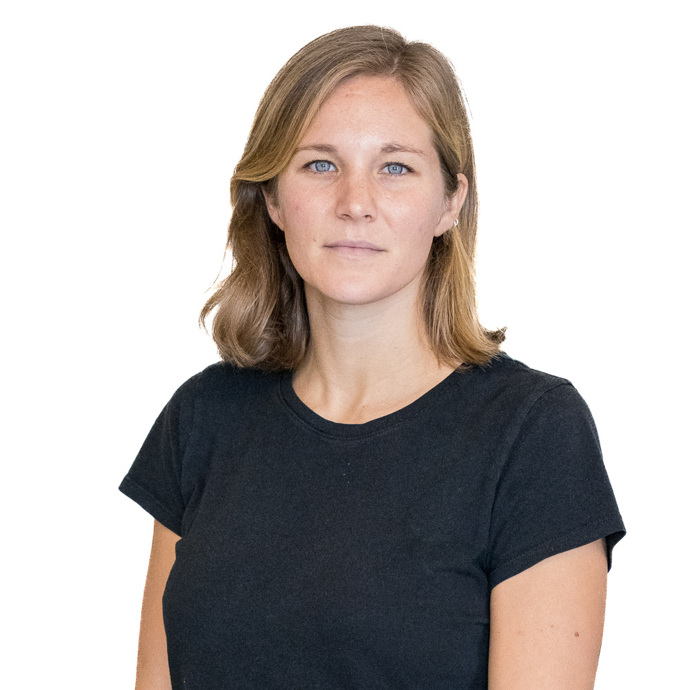 A person with shoulder-length hair and a black t-shirt stands in front of a perforated metal background, looking at the camera with a neutral expression.