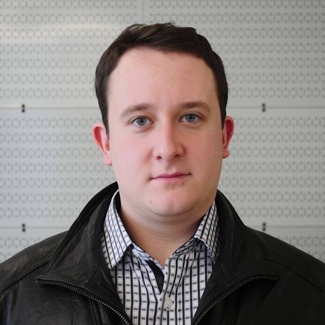 A person with short dark hair is wearing a black leather jacket over a black and white checkered shirt. The background features a wall with a grid pattern.