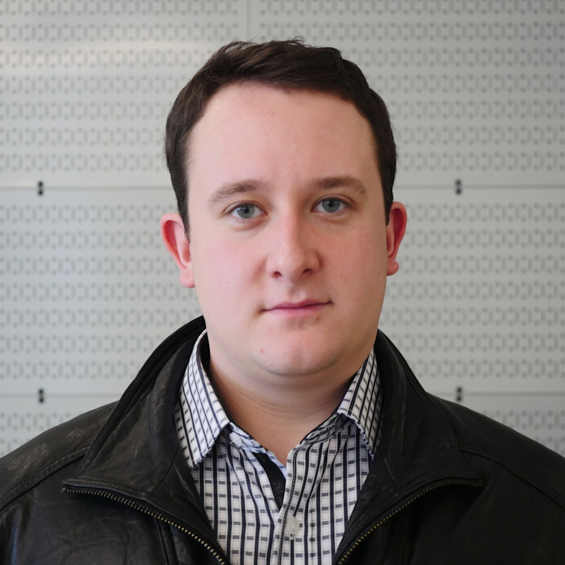 A person with short dark hair is wearing a black leather jacket over a black and white checkered shirt. The background features a wall with a grid pattern.
