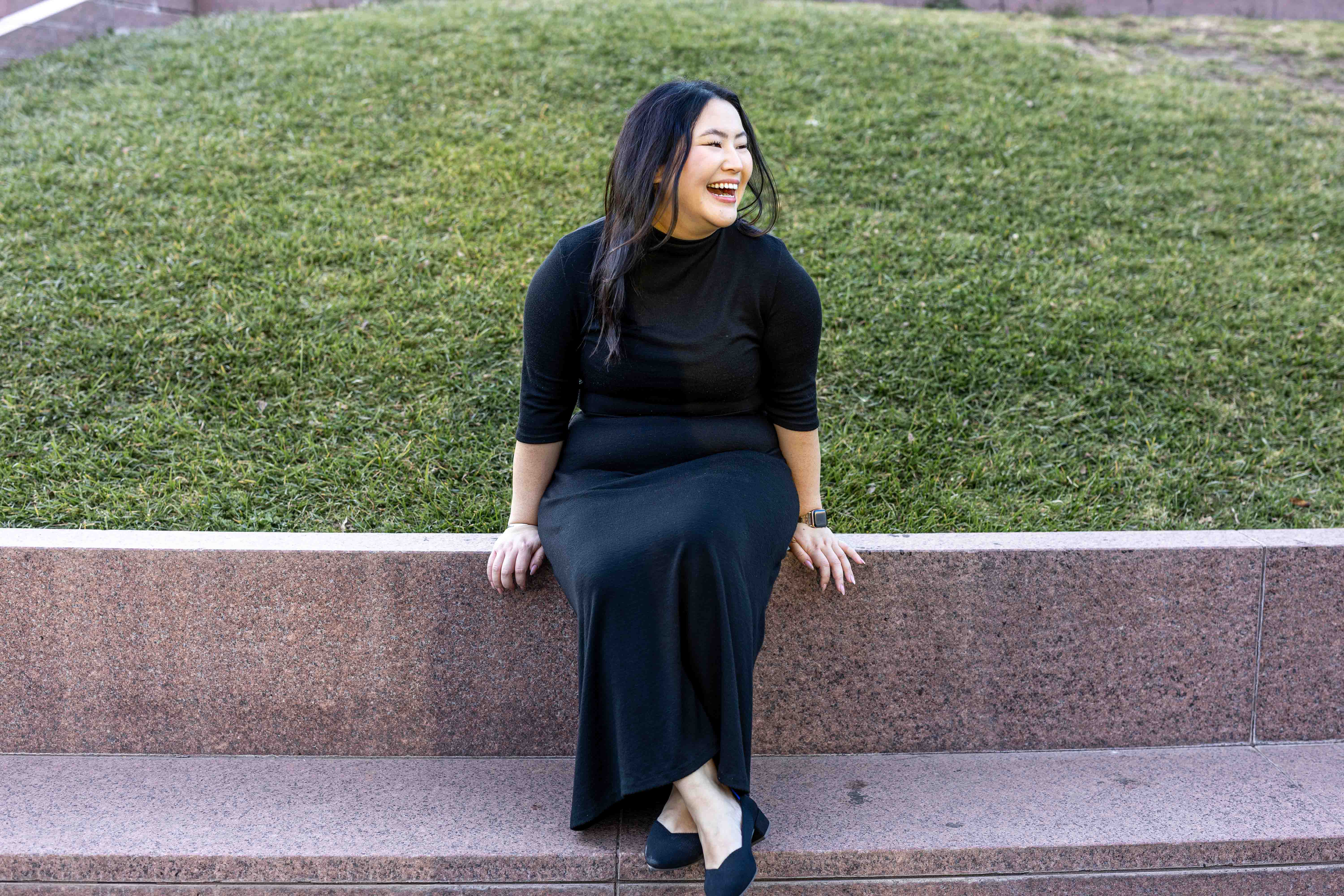 A woman in a black dress and black shoes is sitting on a stone ledge, smiling and looking to the side. She is in front of a grassy hill.