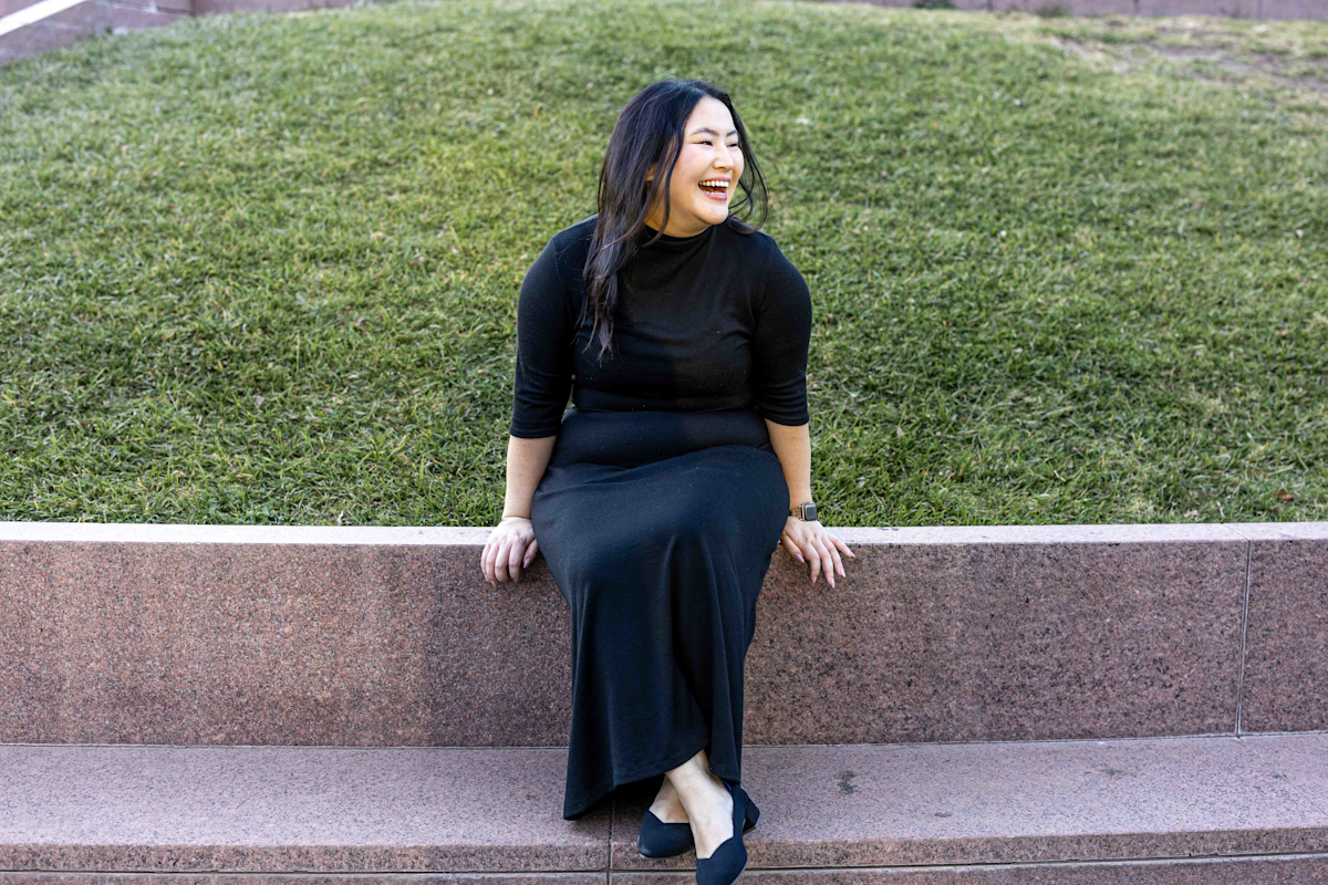 A woman in a black dress and black shoes is sitting on a stone ledge, smiling and looking to the side. She is in front of a grassy hill.