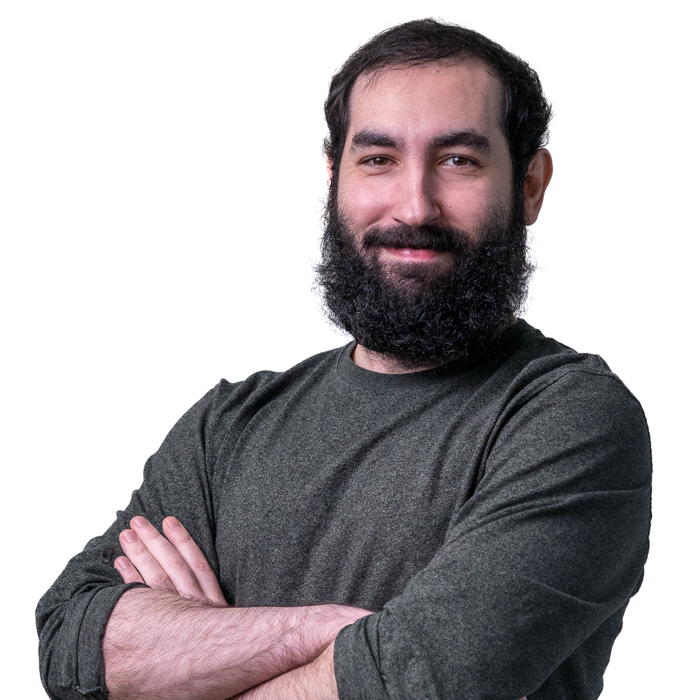 A man with a beard and dark hair, wearing a green sweater, stands smiling with his arms crossed. He is in front of a patterned wall with square perforations.