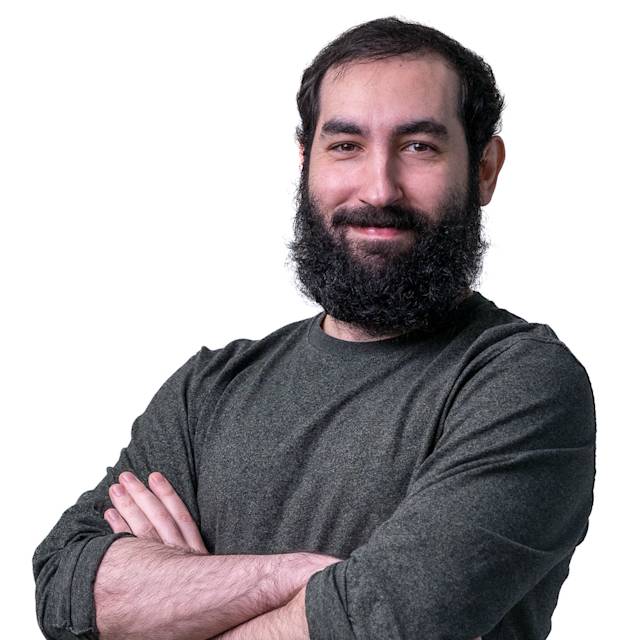 A man with a beard and dark hair, wearing a green sweater, stands smiling with his arms crossed. He is in front of a patterned wall with square perforations.