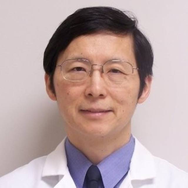A man with short black hair, wearing glasses, a blue collared shirt, and a white lab coat, stands against a plain background. He is facing the camera and smiling slightly.